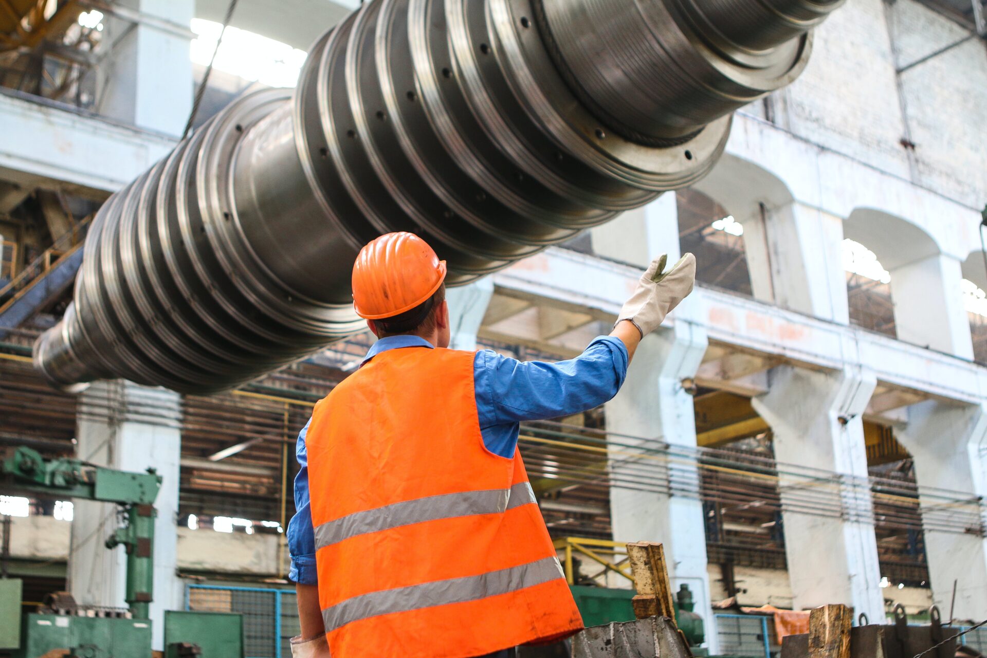 A factory worker in safety gear gestures towards large industrial machinery, representing the robust work ethic in Europe's manufacturing sector.